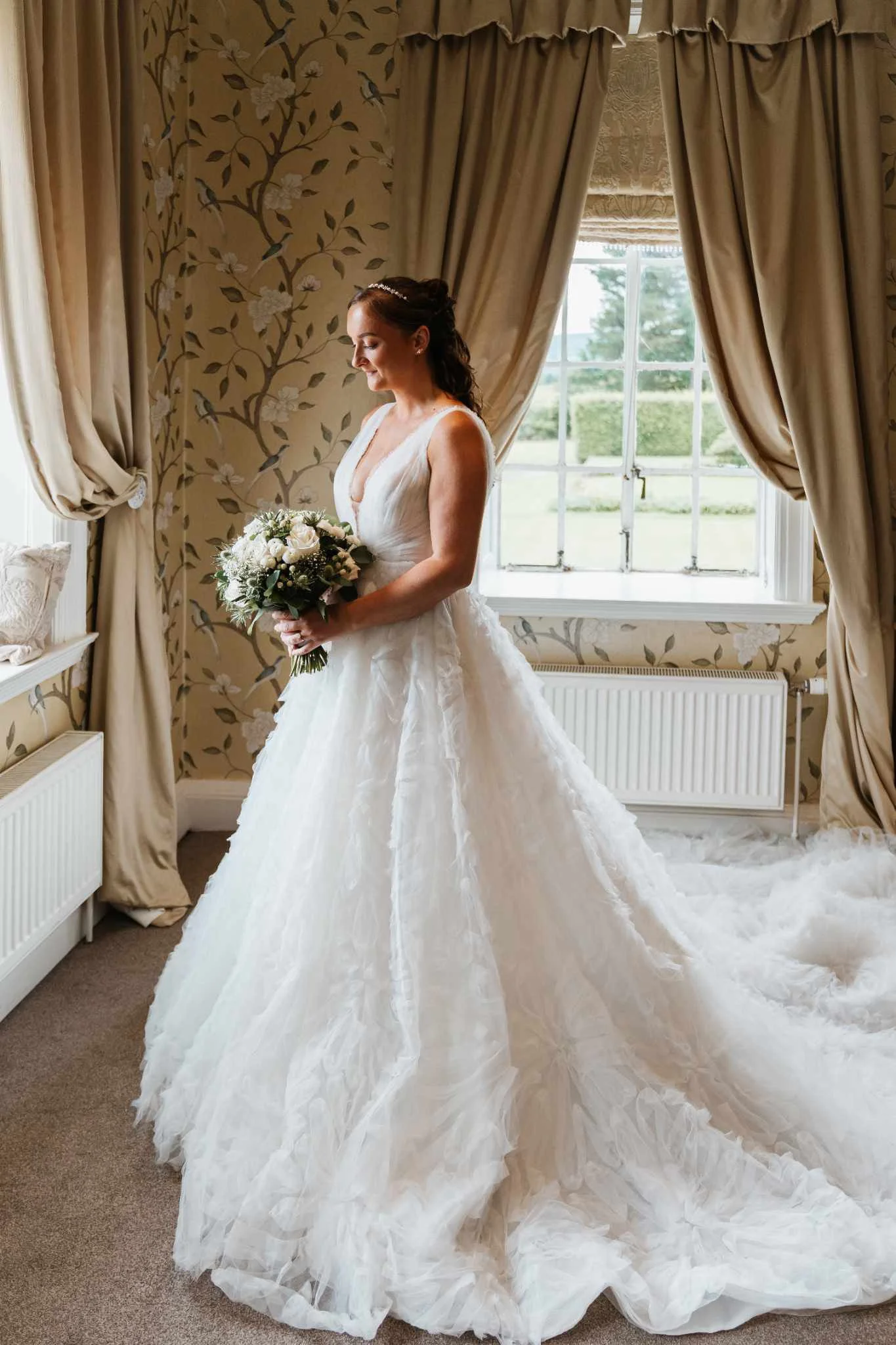 A bride in a white wedding gown holding a bouquet of flowers, standing in a room with floral wallpaper and beige curtains, near a window.