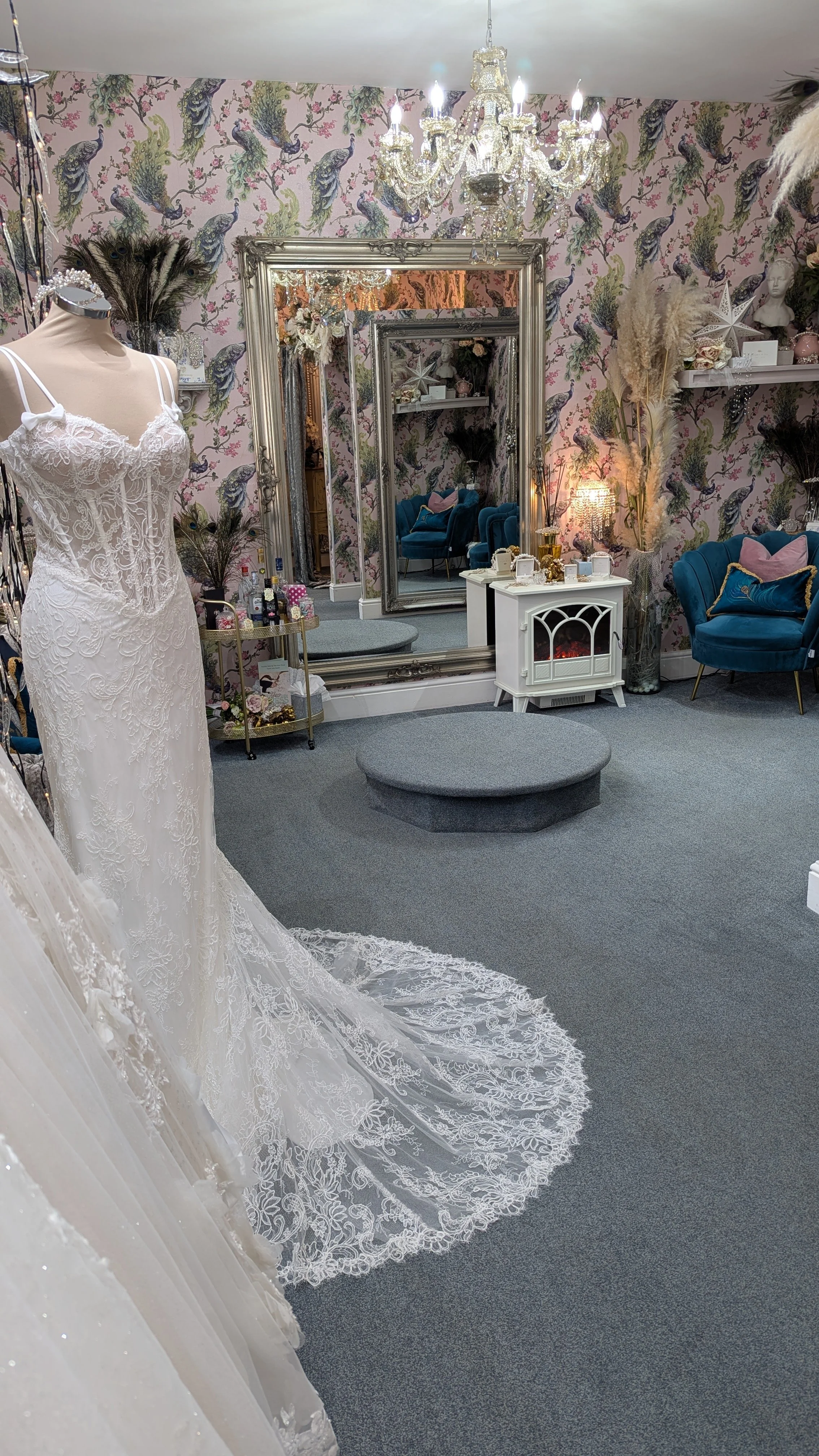 Interior of a bridal boutique with a white lace wedding dress on display, large ornate mirror, plush blue chairs, decorative wallpaper with peacocks, and elegant decor including a chandelier, pampas grass, and a small fireplace.