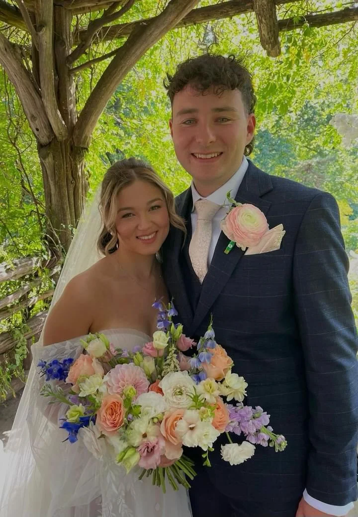 A bride and groom smiling for a wedding photo outdoors under a tree, with the bride holding a large bouquet of colorful flowers.