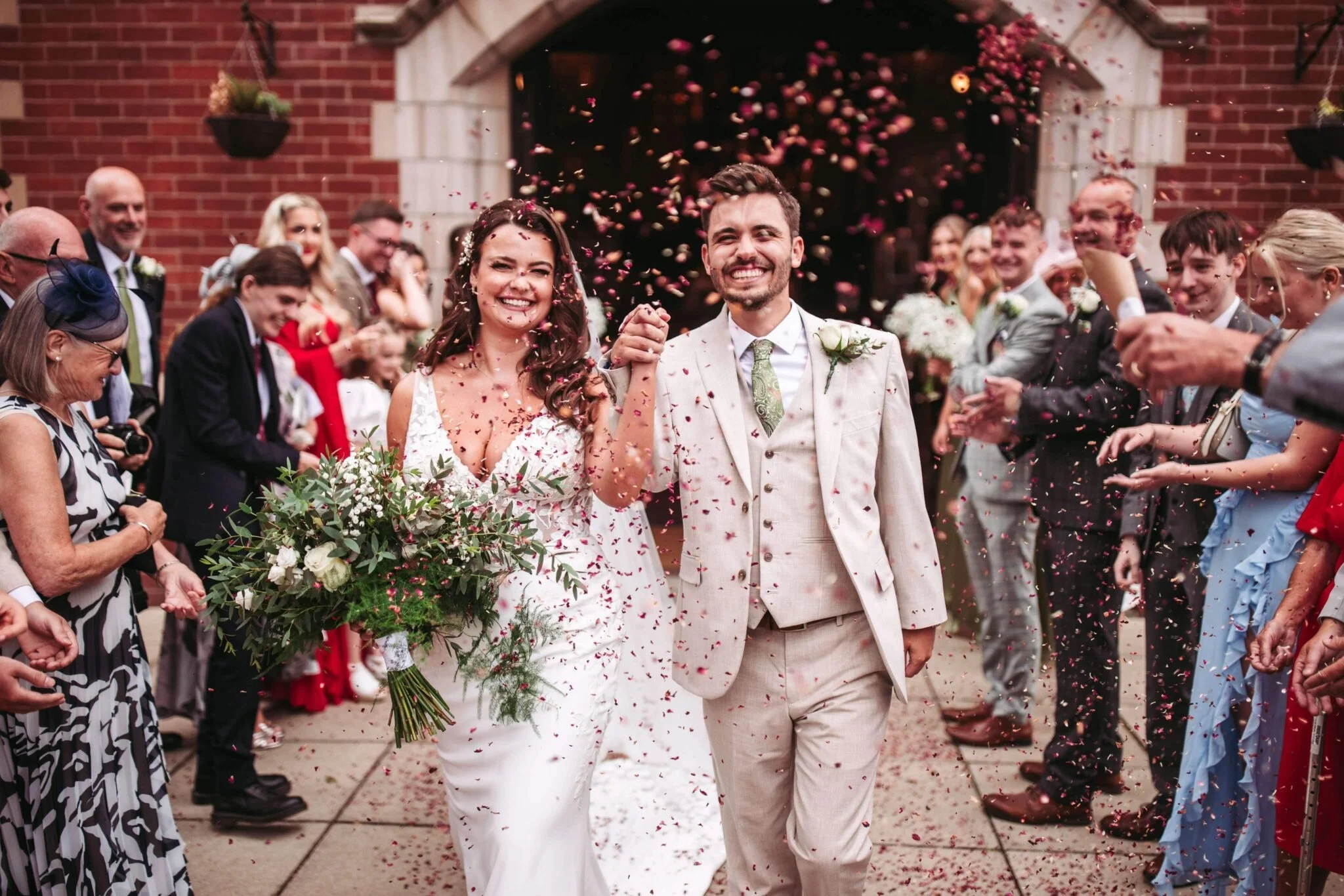 A recently married couple smiles joyfully as they walk hand-in-hand through a crowd of friends and family, with pink confetti falling around them outside a building with brick walls.