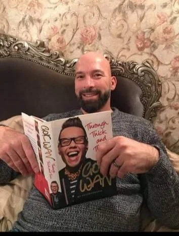 Man with a beard smiling and holding a magazine while sitting on a bed with a decorative headboard and floral wallpaper in the background.