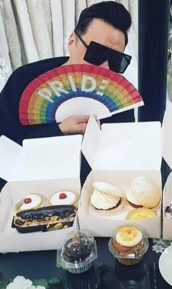 Person wearing sunglasses holding a rainbow-colored fan with the word "PRIDE" on it. In front of them are opened white boxes with donuts, including one with a cherry on top, and some drinks.