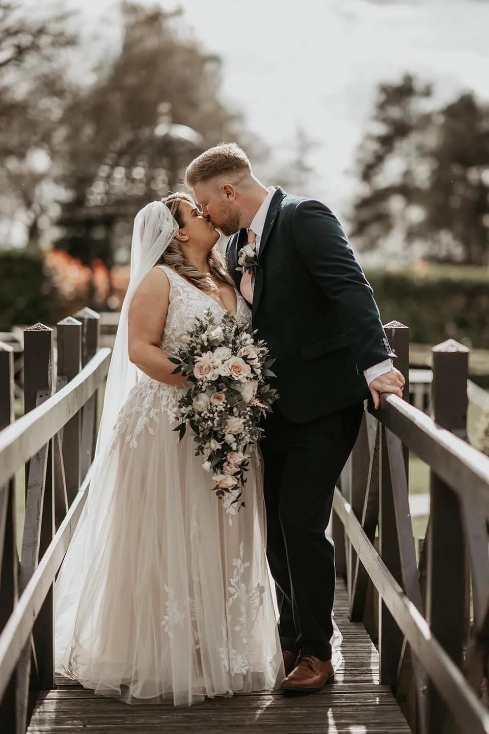 A bride and groom lean in for a kiss on a wooden bridge outdoors, holding a bouquet with roses and greenery.