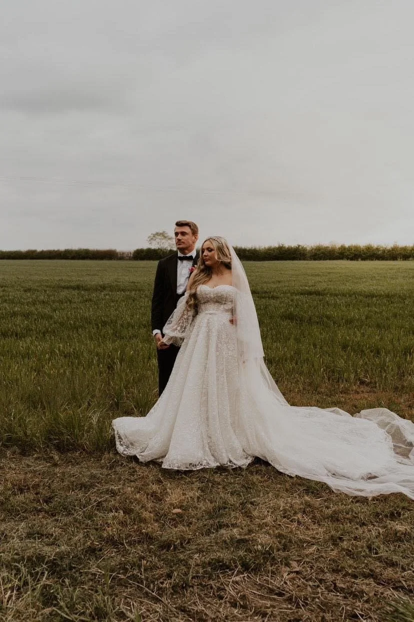 A bride and groom standing hand in hand in an open field, with the bride wearing a long white wedding gown and the groom in a black tuxedo and bow tie, under a cloudy sky.