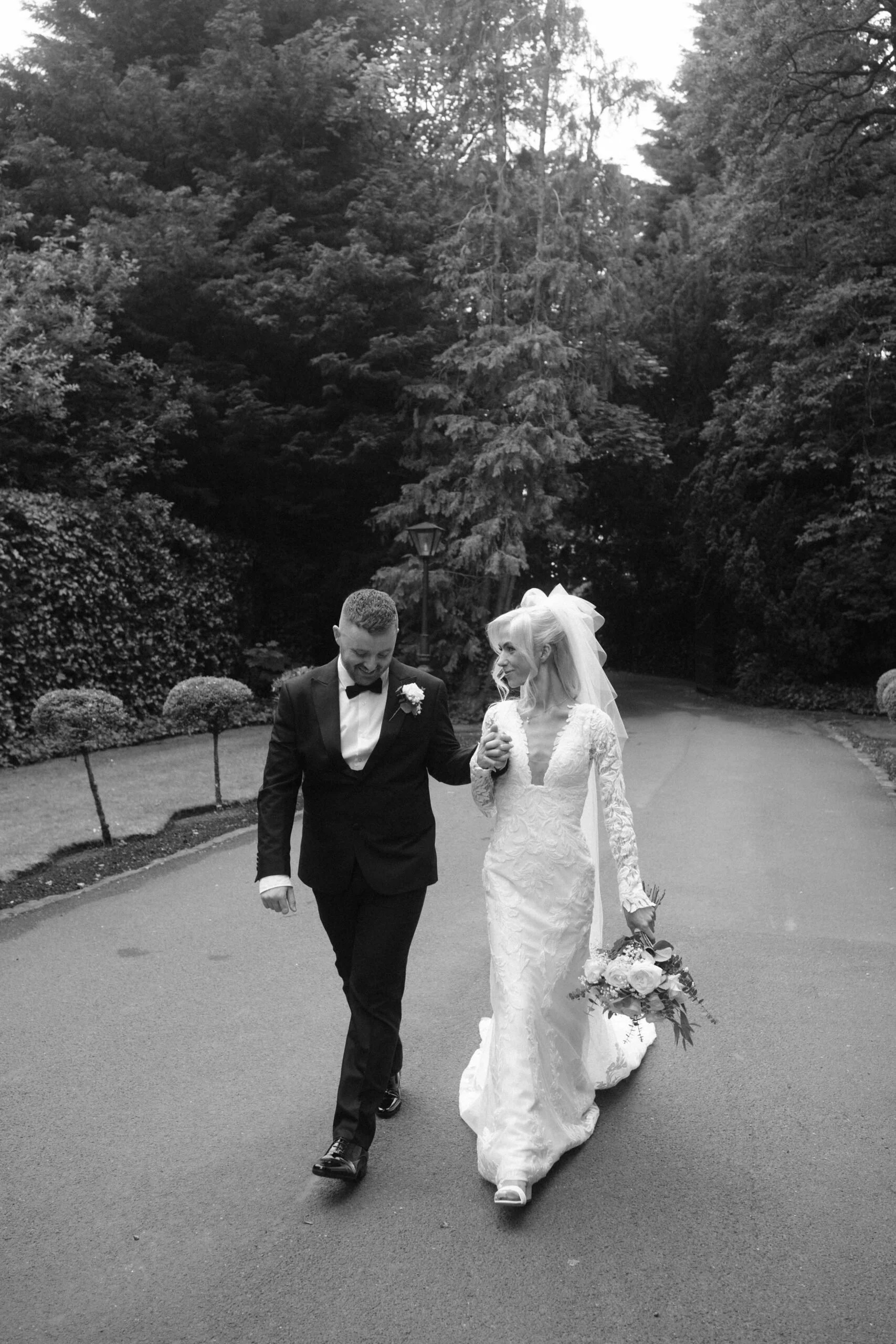 A bride and groom walking hand in hand outdoors on a paved path, surrounded by trees and bushes, on their wedding day.