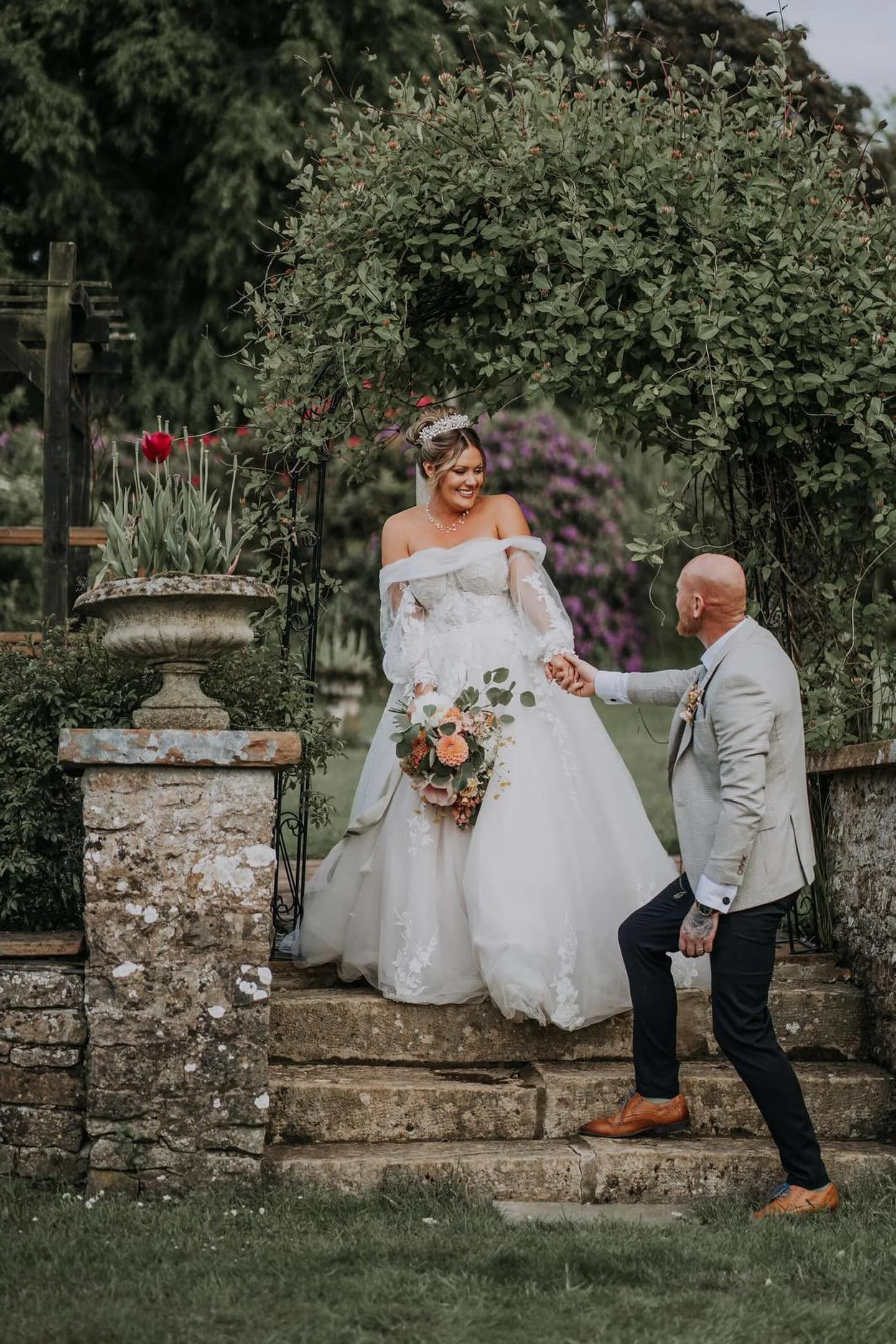 A bride in a white wedding gown and a man in a light gray suit holding hands on outdoor stone steps, surrounded by greenery and flowers.