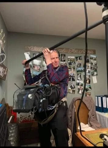 A man holding a boom microphone in a room with photo collage on the wall, laptop, and office supplies.