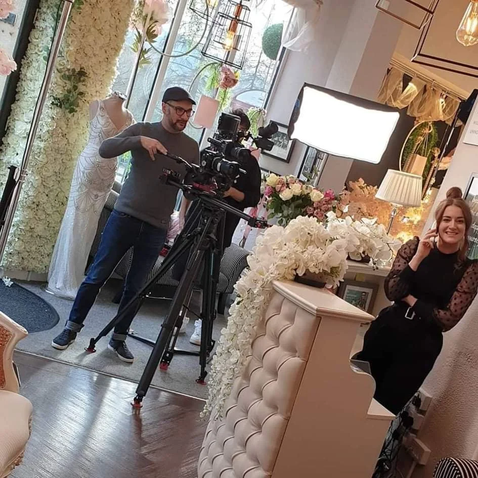 A woman smiling and talking on the phone inside a boutique or flower shop with a photographer filming her. The scene includes floral arrangements and mannequins.