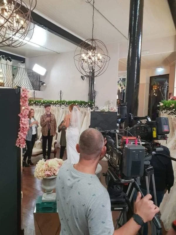 Wedding ceremony underway with bride in white gown and veil, surrounded by guests, at a decorated indoor venue with floral arrangements and chandelier lighting.