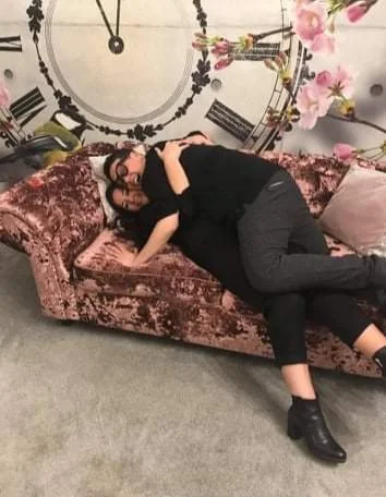A woman lying on a vintage pink velvet sofa, hugging a pillow, in front of a large decorative wall clock with floral accents.