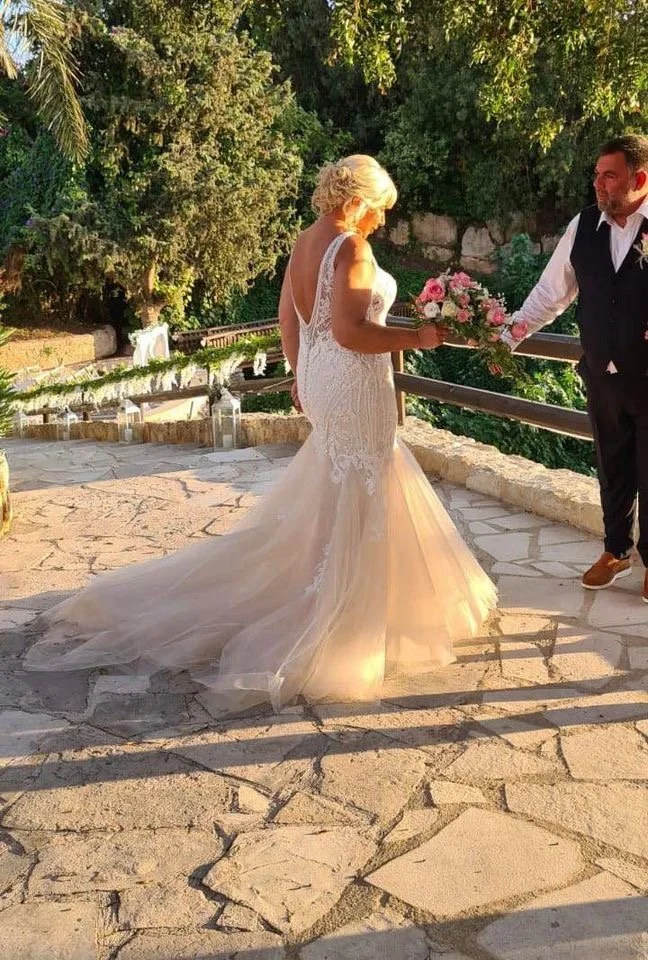 A bride in a white wedding gown with lace details holding a bouquet of pink flowers is receiving flowers from a groom in a black vest and white shirt on an outdoor stone platform with greenery in the background.