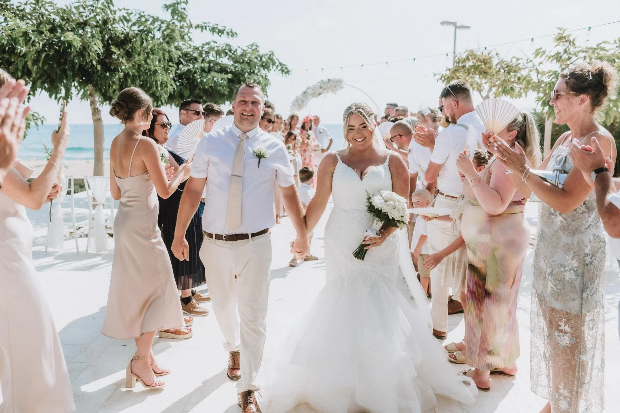 Bride and groom walking hand in hand down aisle at beach wedding, surrounded by smiling guests clapping and taking photos on a sunny day.