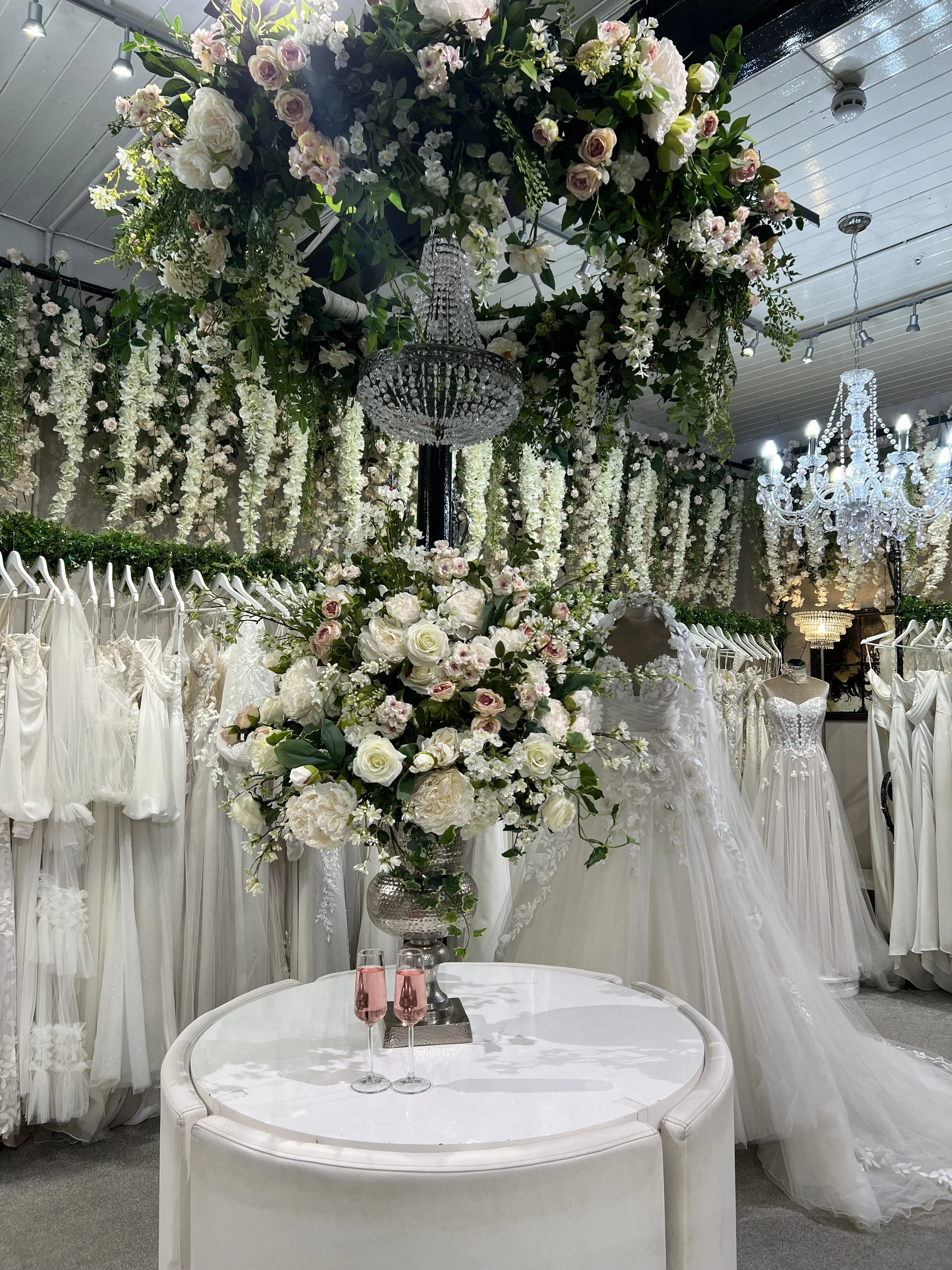 Elegant bridal shop display with wedding dresses, large floral arrangements, chandeliers, and a white table with two pink champagne glasses.