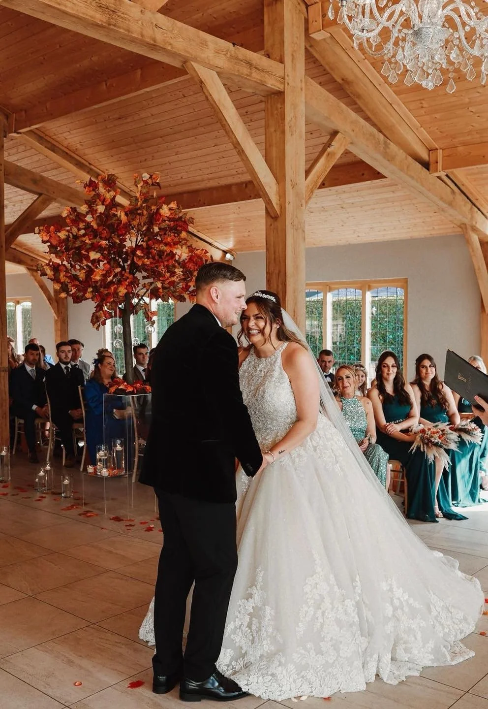 Bride and groom exchanging vows during wedding ceremony in a rustic venue with wood beams, large window, chandelier, and seating for guests.
