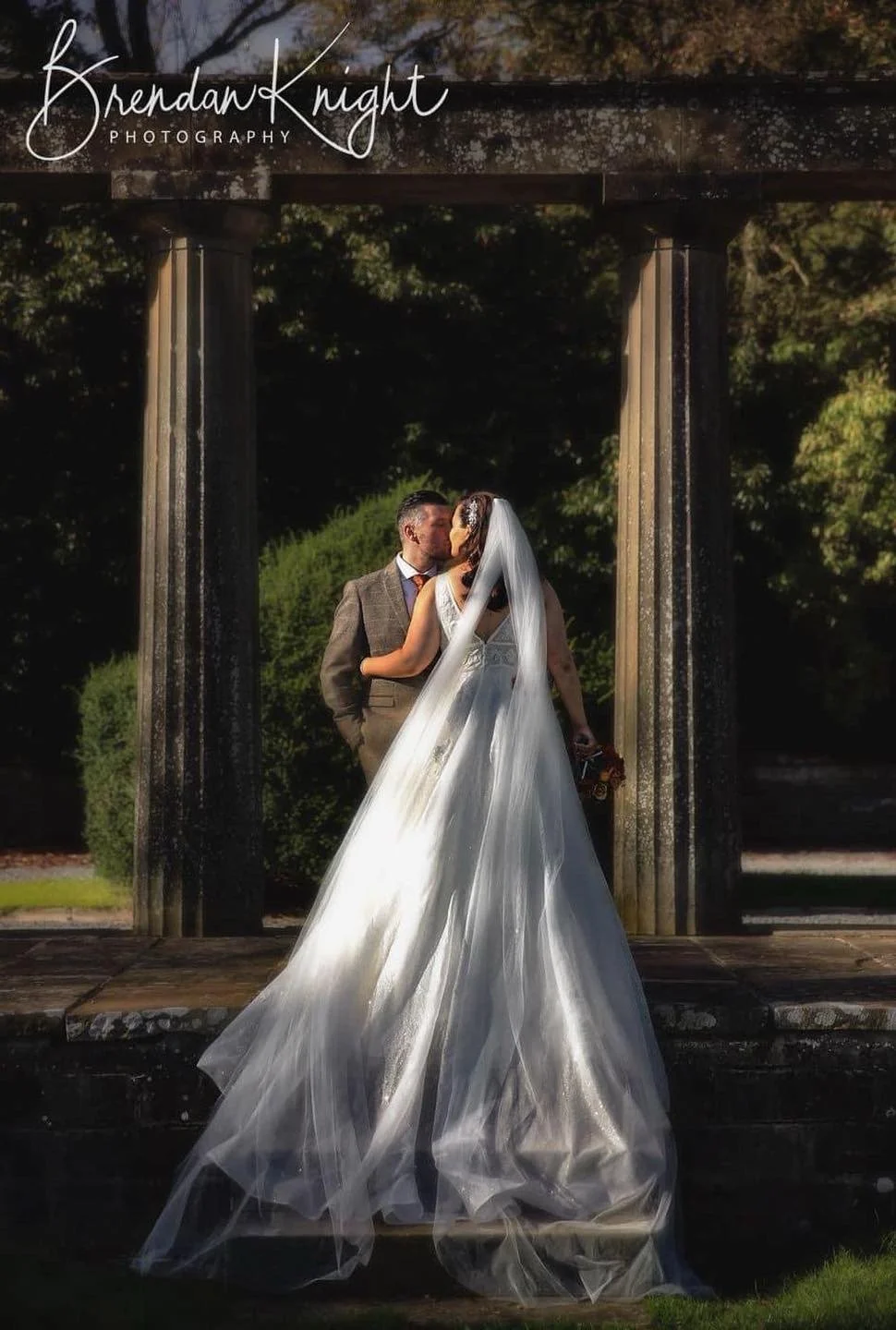 A bride and groom kissing outdoors beneath a stone arch, with the bride wearing a long train and veil, and the groom in a suit, during a wedding photoshoot.