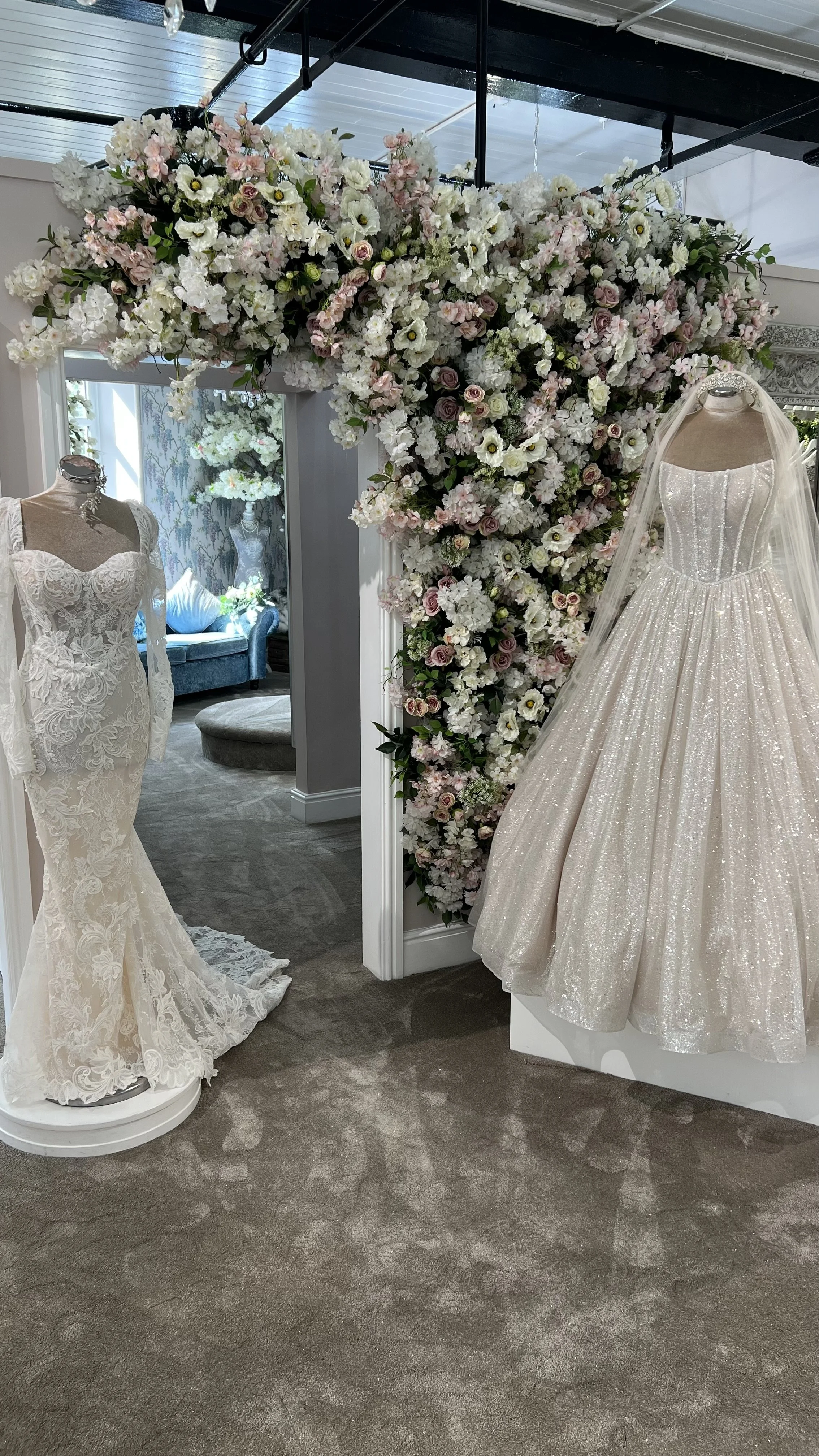 Wedding dress shop display with two mannequins wearing bridal gowns, one with lace detailing and the other with a glittery fabric, amid a floral arch of pink, white, and green flowers.