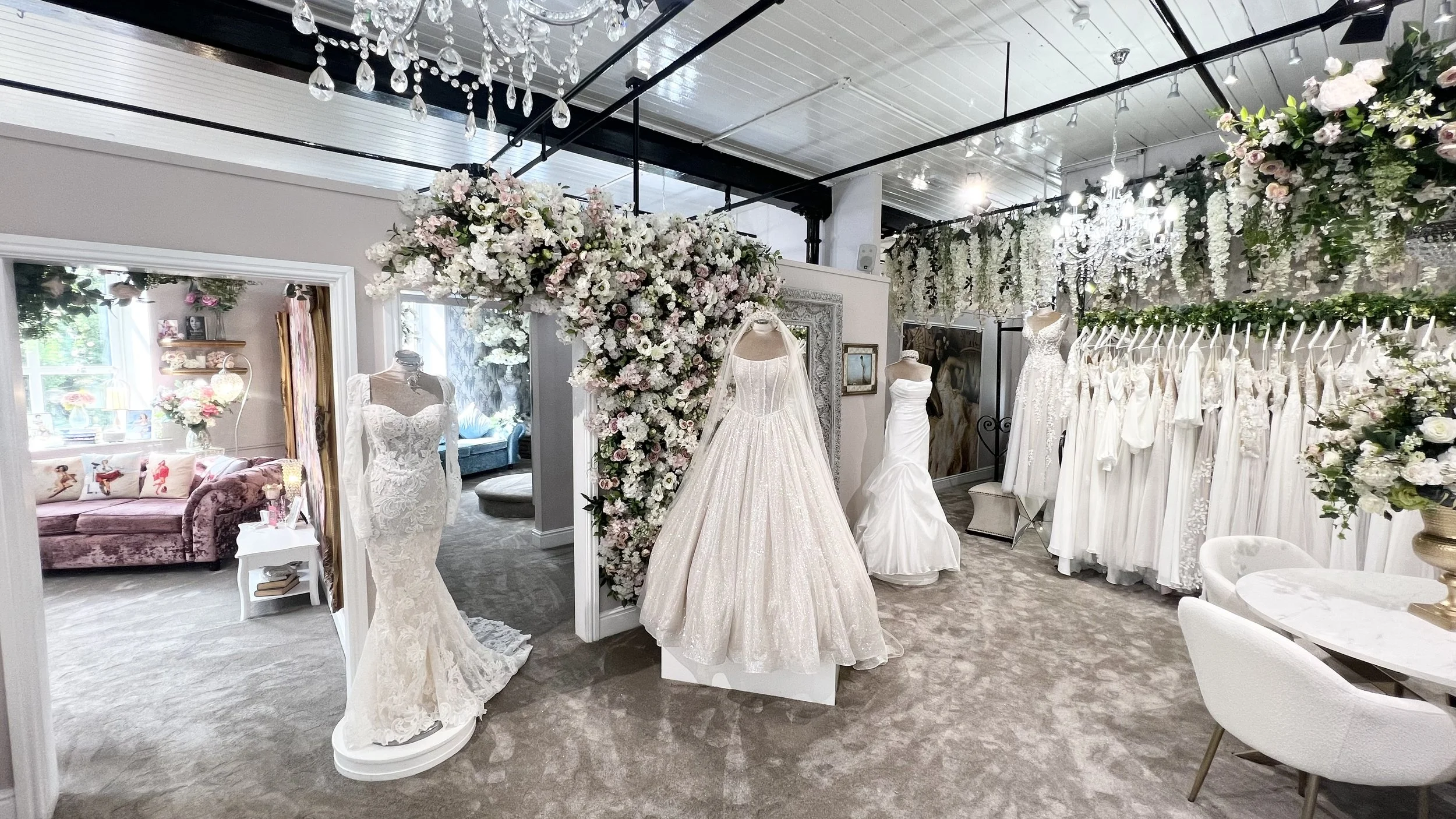 The interior of a bridal boutique with wedding dresses on display, decorated with floral arrangements and chandeliers.