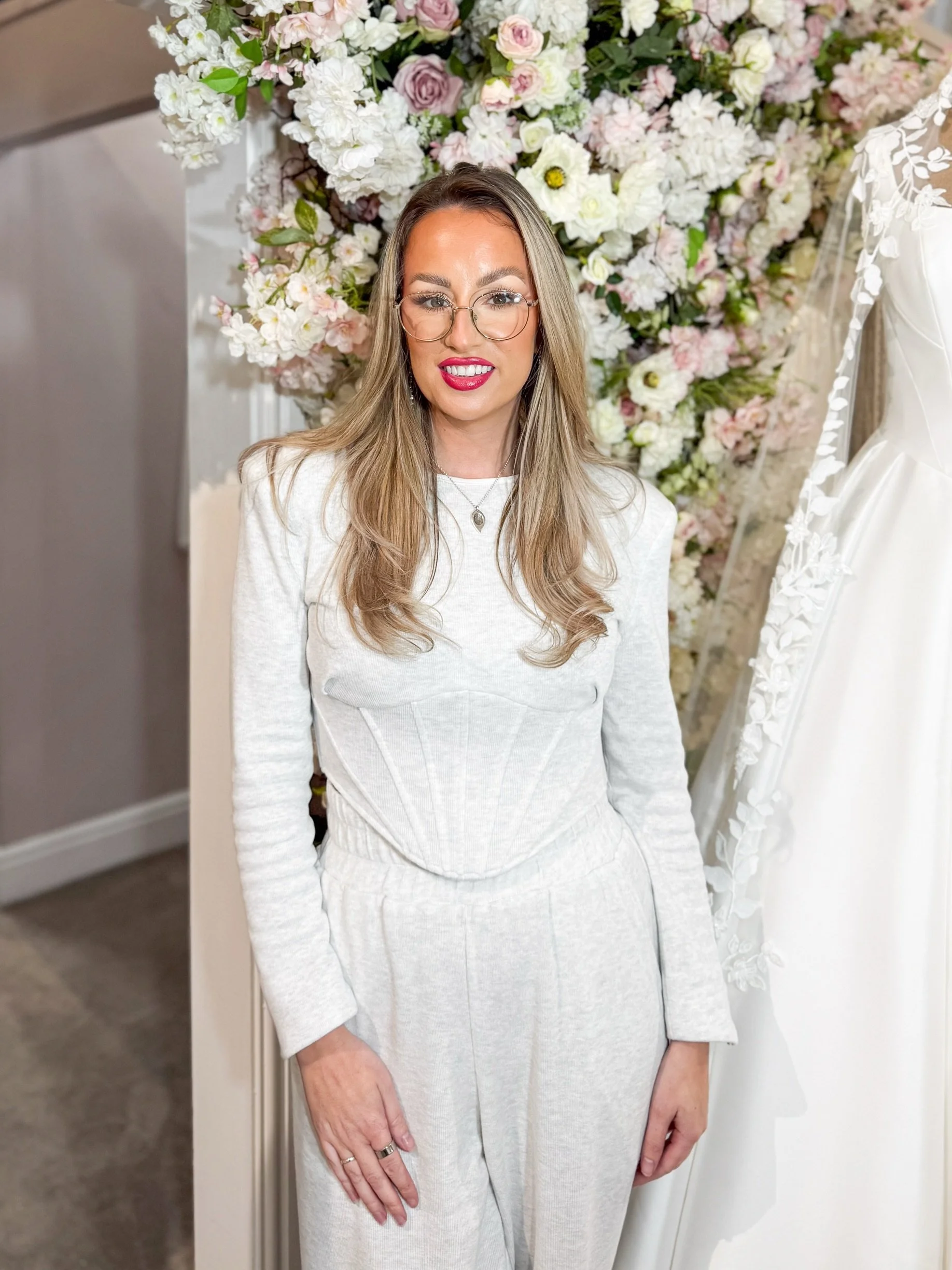 A woman with long blonde hair, glasses, and a big smile, stands in a bridal shop wearing a white sleeveless zip-up top and white pants. She is surrounded by wedding dresses on racks and a mannequin displaying a wedding gown.