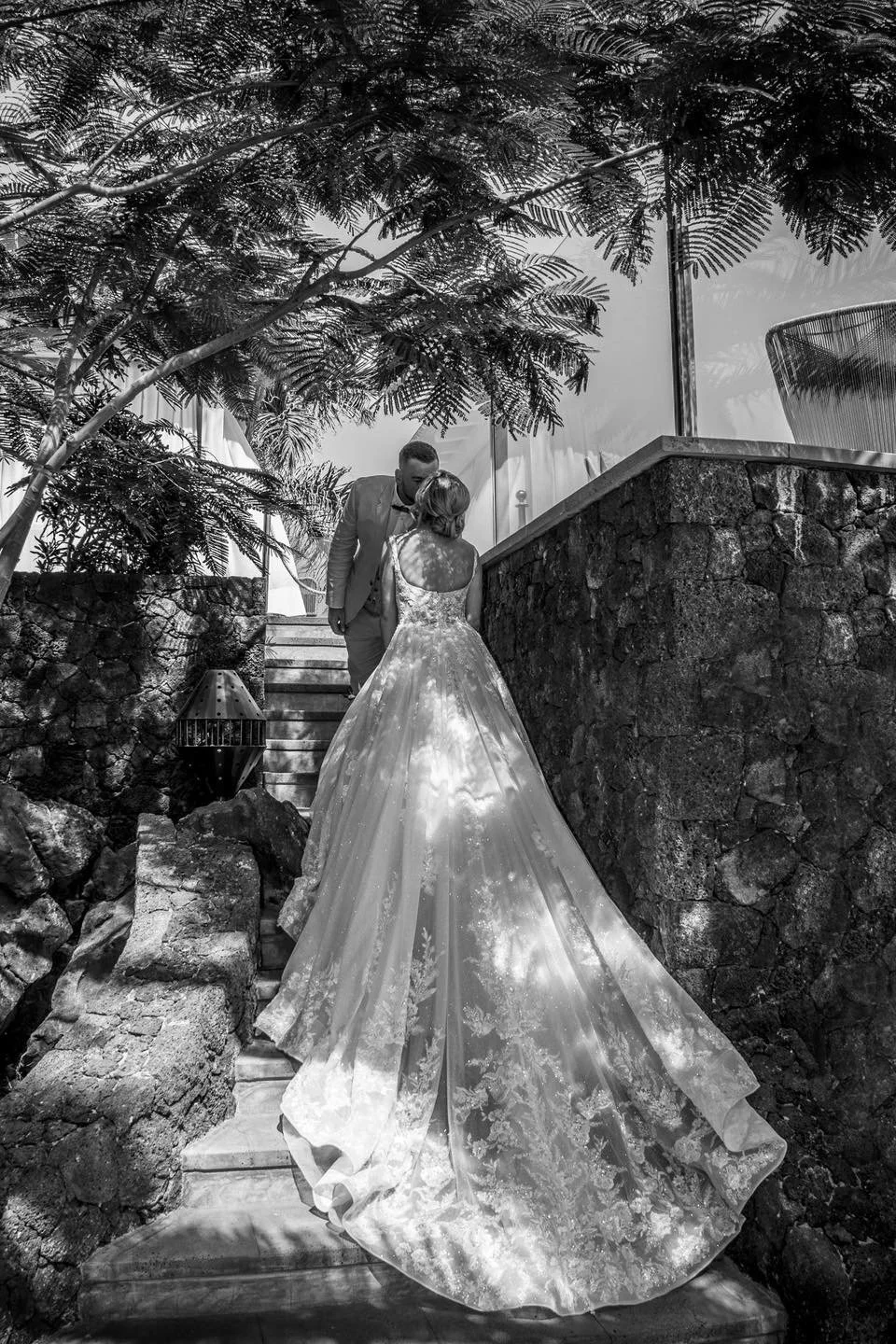 A bride and groom share a kiss on stone steps, surrounded by trees, in a black and white wedding photo.