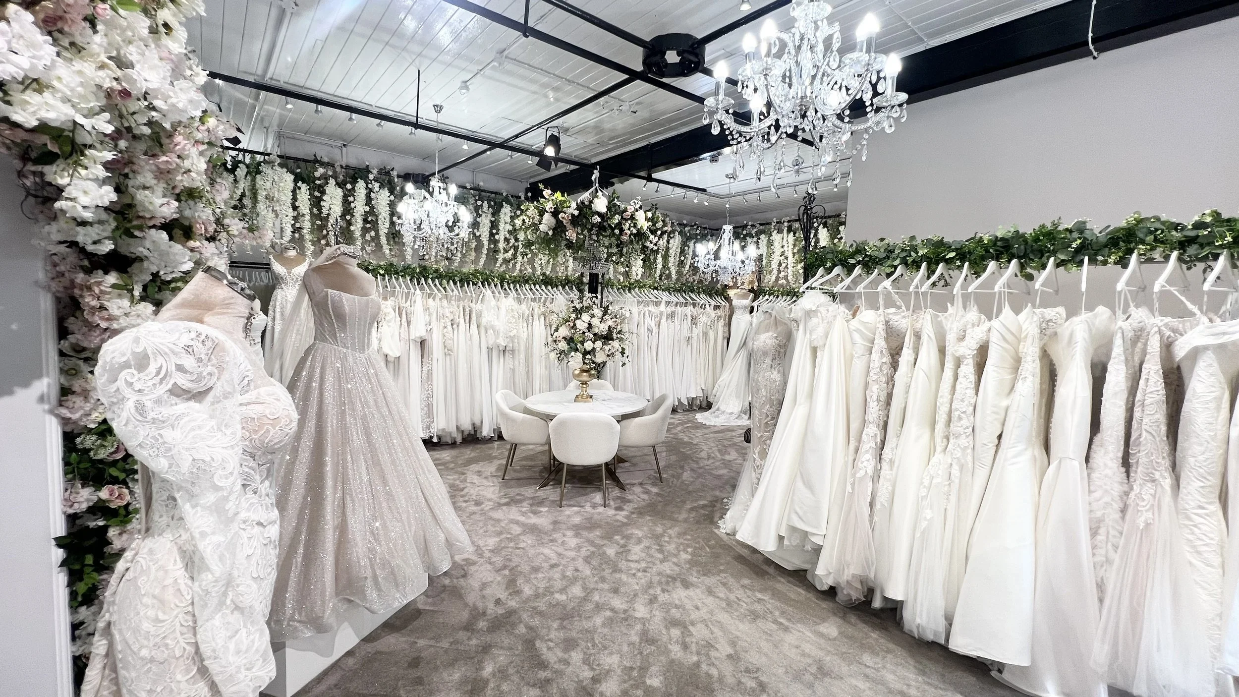 A bridal boutique filled with elegant wedding dresses on display, floral decorations, chandeliers, and a round table with chairs in the center, creating a luxurious shopping environment.