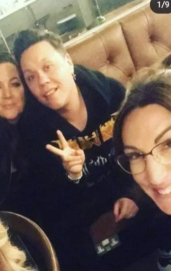 Group of three women smiling and taking a selfie in a restaurant or cafe with a brown leather booth and kitchen appliances in the background.