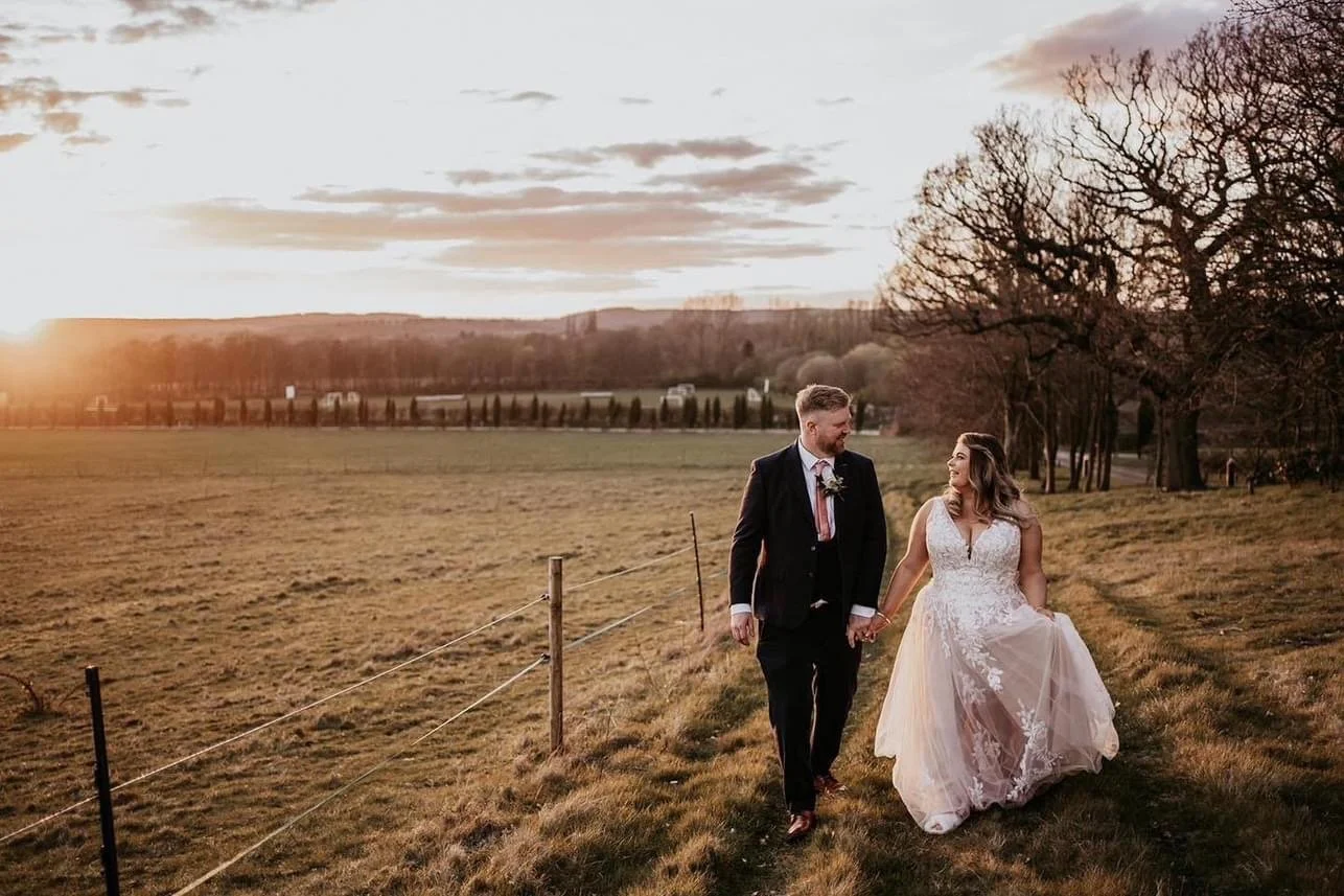 A bride and groom walking hand in hand outdoors at sunset, with a field and trees in the background.