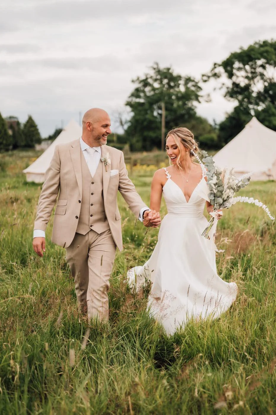 A bride and groom walking hand in hand through a grassy field on their wedding day, with tents and trees in the background.