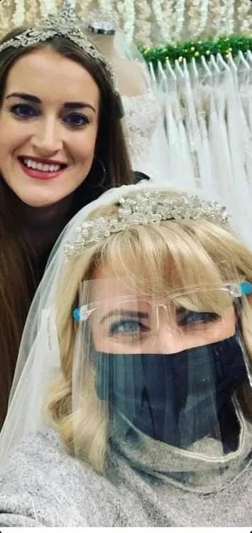 Two women, one smiling and wearing a crown and the other wearing a face shield and mask, taking a selfie at a celebration or event with white curtains and greenery in the background.