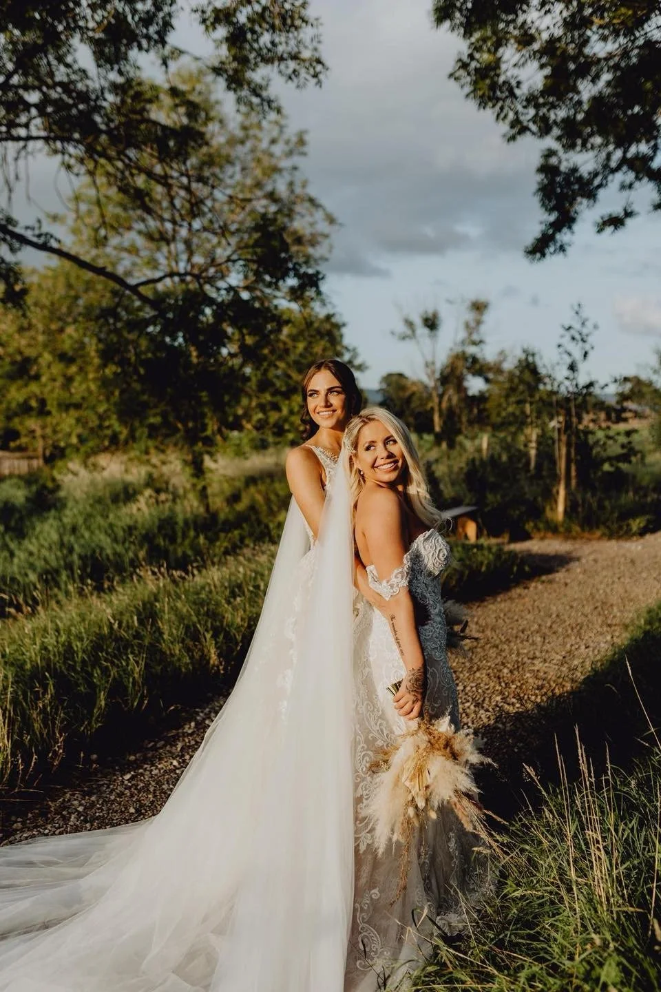 Two women in wedding dresses, smiling and posing outdoors among trees and grass during sunset.