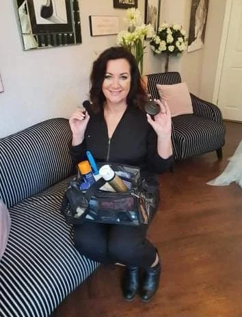 A woman sitting on a striped black and white sofa, holding a makeup powder compact and a makeup brush, with a makeup bag and beauty products in her lap, smiling at the camera.