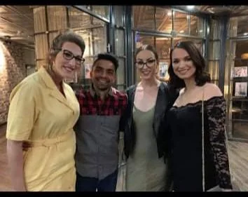 Four people smiling and posing together indoors, with a glass wall and metal framework in the background.