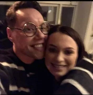 A man and a woman smiling closely together in a selfie, indoors.