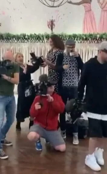 Group of people in a room, some are taking photos or videos, others are talking or standing around. One person is kneeling with a camera in front of them.