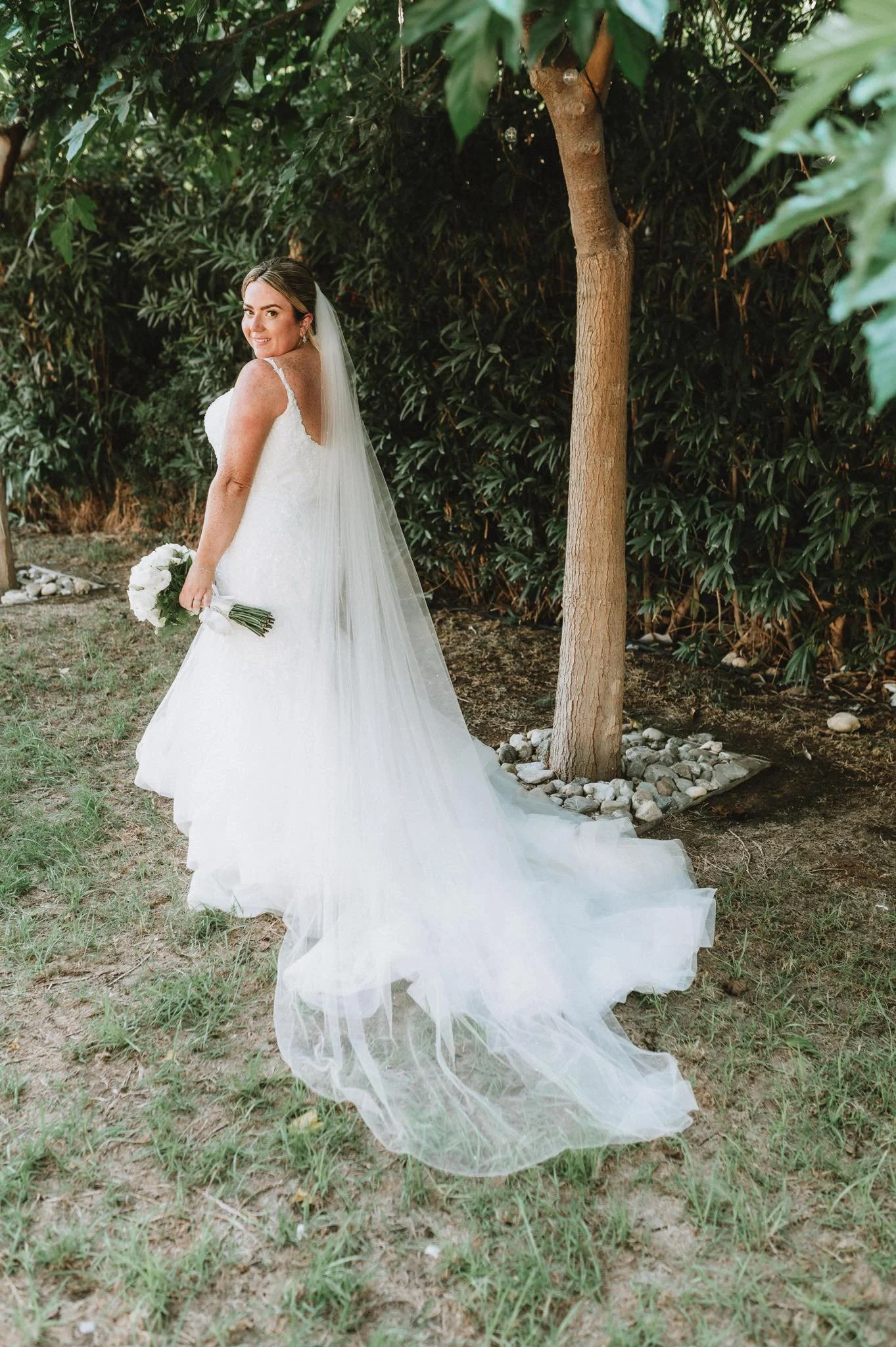 A bride in a white wedding gown holding a bouquet of white flowers, standing outdoors next to a tree and dense green foliage.