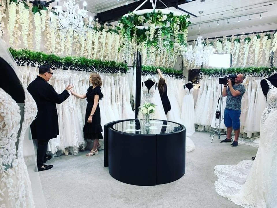People shopping for wedding dresses in a bridal boutique, with racks of white wedding gowns and floral decorations.