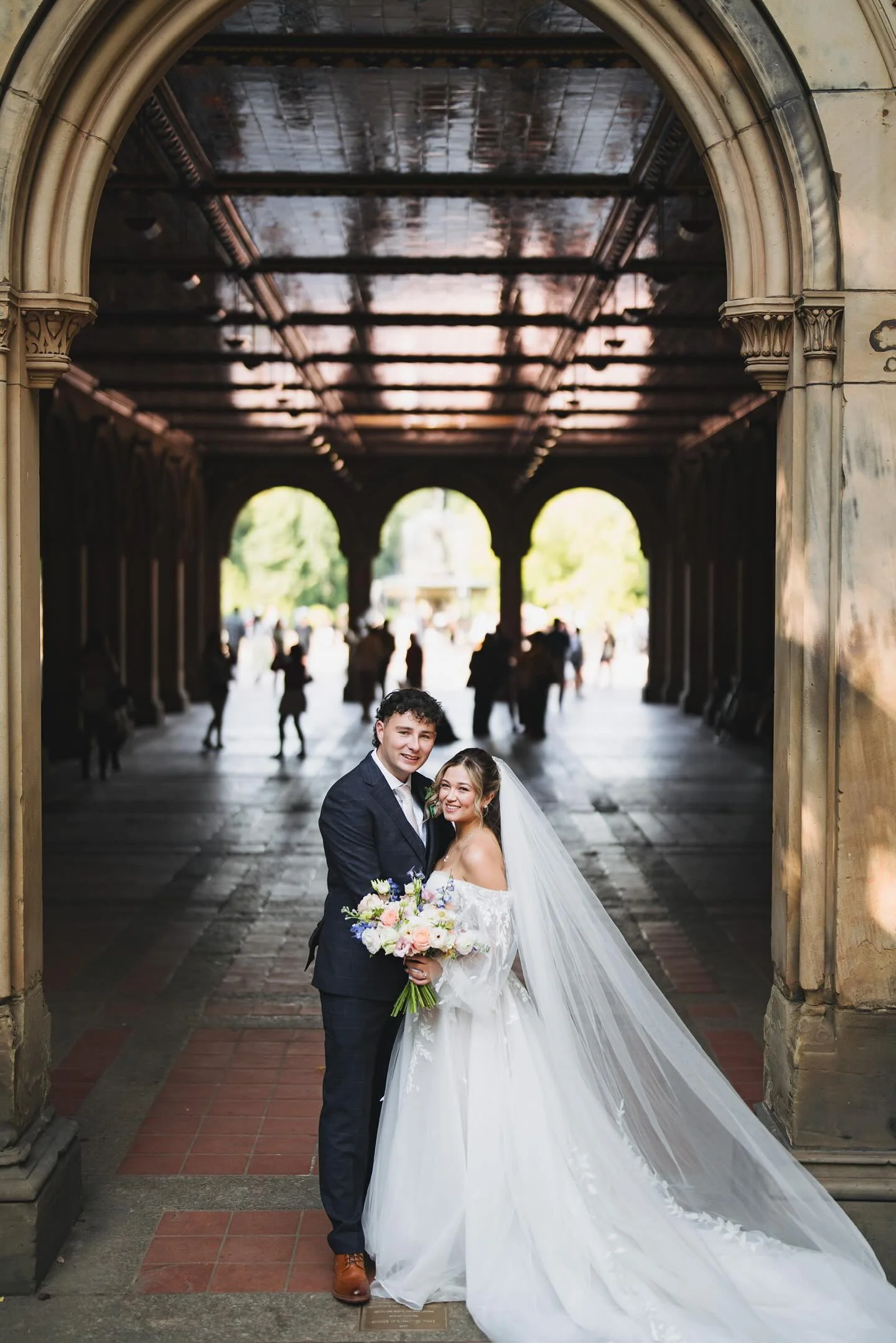 A newlywed couple stands under an archway, smiling at the camera. The bride is wearing a white wedding dress with a long train and veil, holding a bouquet of flowers. The groom is in a dark suit with a white shirt. People are visible in the backgroun