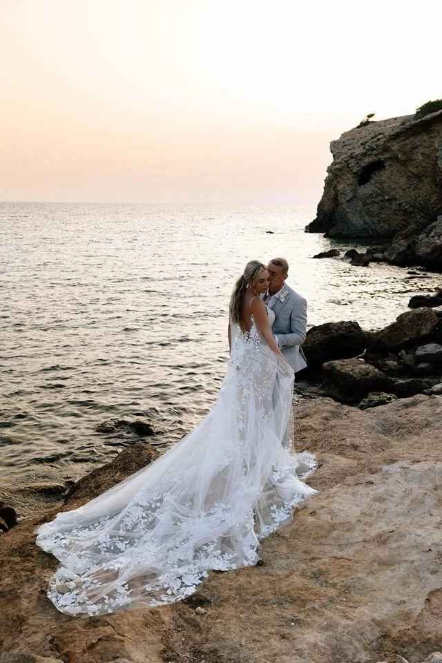 A bride in a white wedding gown and a groom in a light gray suit standing close together on a rocky shoreline during sunset, near the ocean.