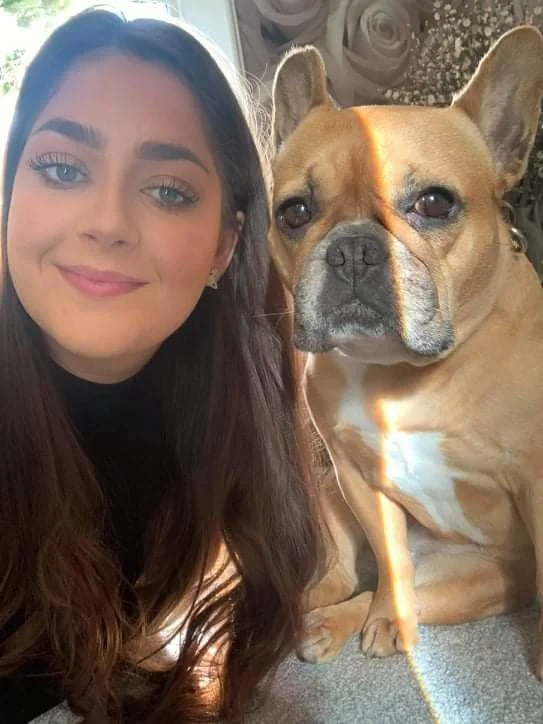 A young woman with long dark hair and blue eyes taking a selfie with a tan French Bulldog with a black nose and dark eyes.