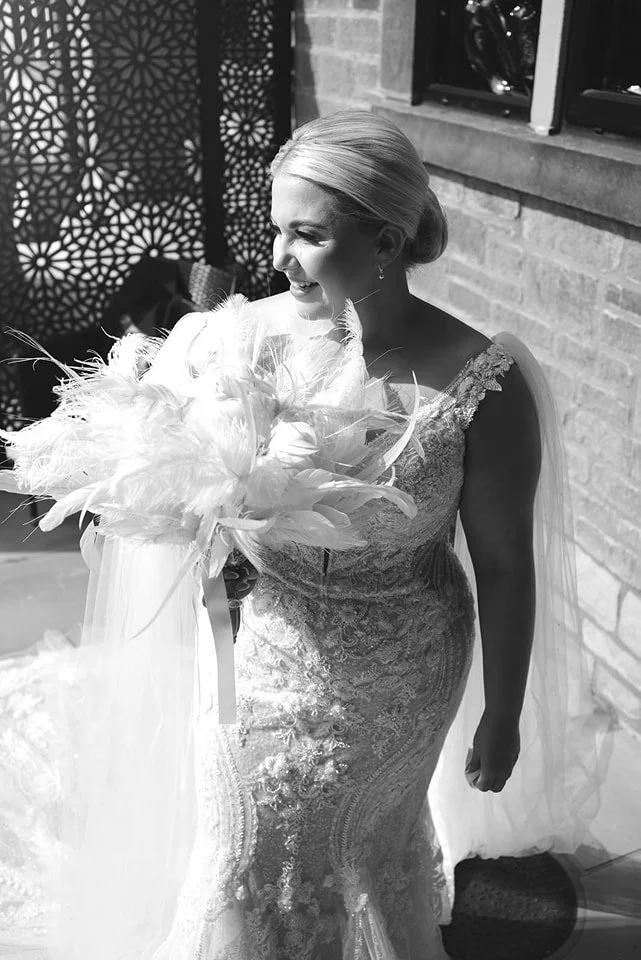A bride with a sleek updo hairstyle, holding a bouquet of feathers, smiling inside a room with brick walls and decorative lattice paneling.