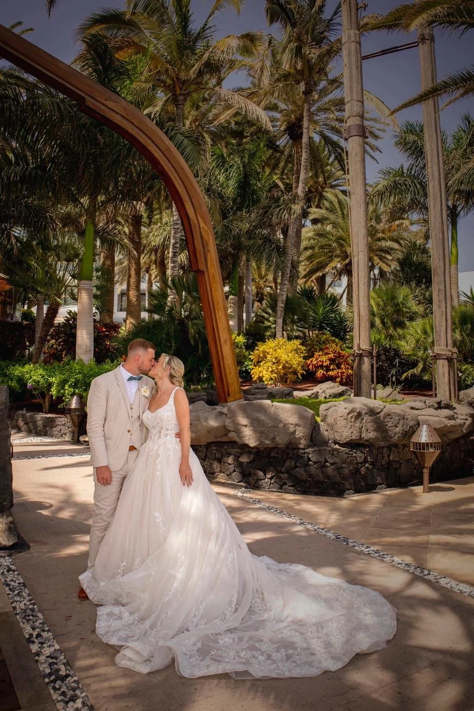 A bride and groom kiss outdoors under a large wooden arch, surrounded by tropical palm trees and lush greenery on a sunny day.
