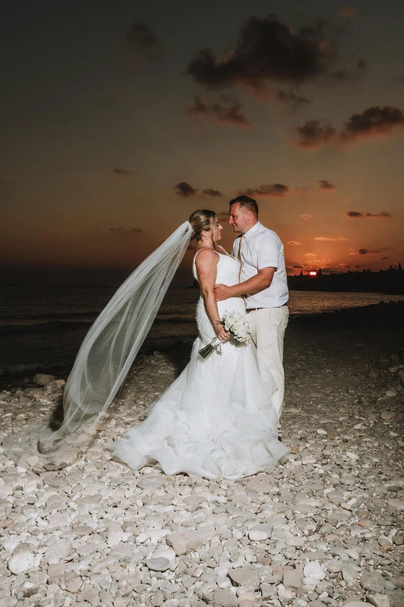 A bride and groom stand close together on a rocky beach at sunset, gazing at each other, with the sky showing a colorful sunset background.