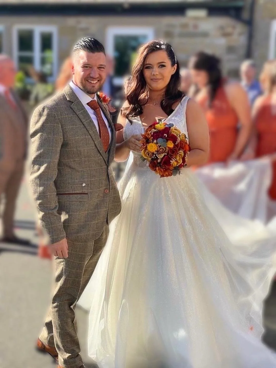 A bride and groom standing outdoors during a wedding celebration, with the bride holding a bouquet of flowers and wearing a white gown, and the groom in a brown plaid suit. Bridesmaids dressed in orange are blurred in the background.