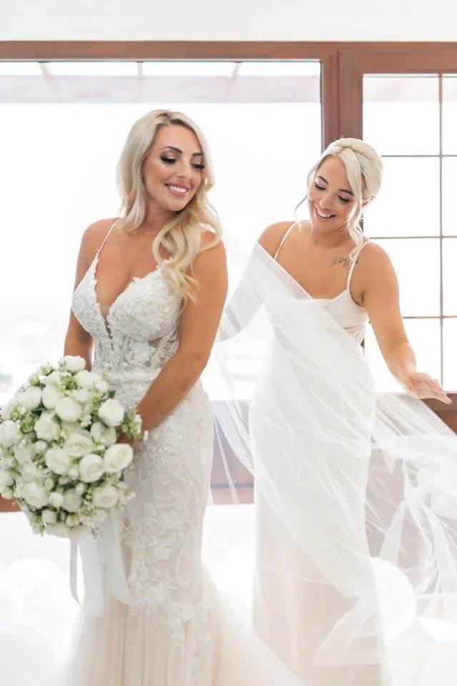 Two women, one in a wedding dress holding a bouquet of white flowers, and another in a white gown helping with the veil, standing near windows in a bright room.