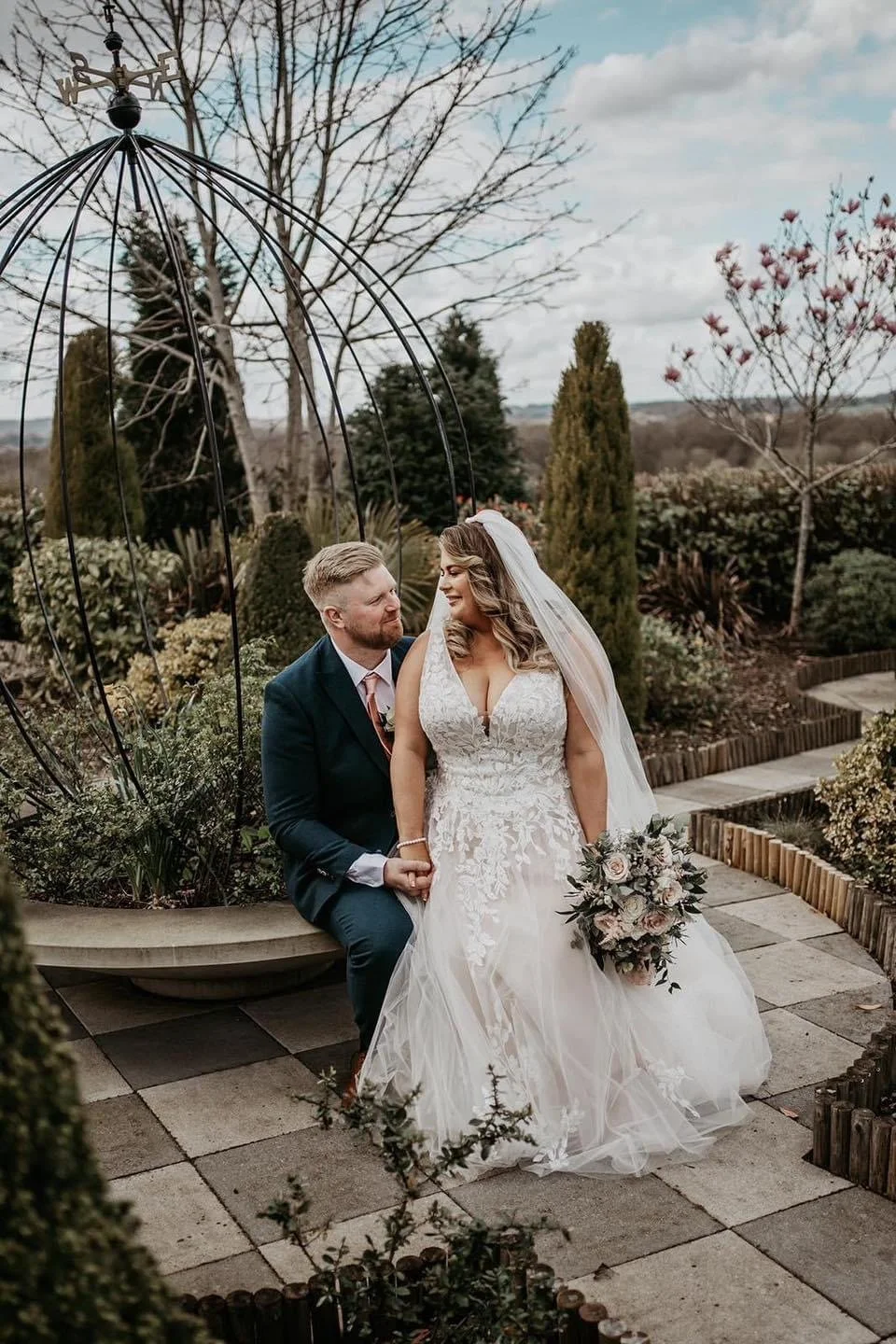 A bride and groom sitting together in a garden, holding hands, with the bride holding a bouquet, surrounded by trees and plants.