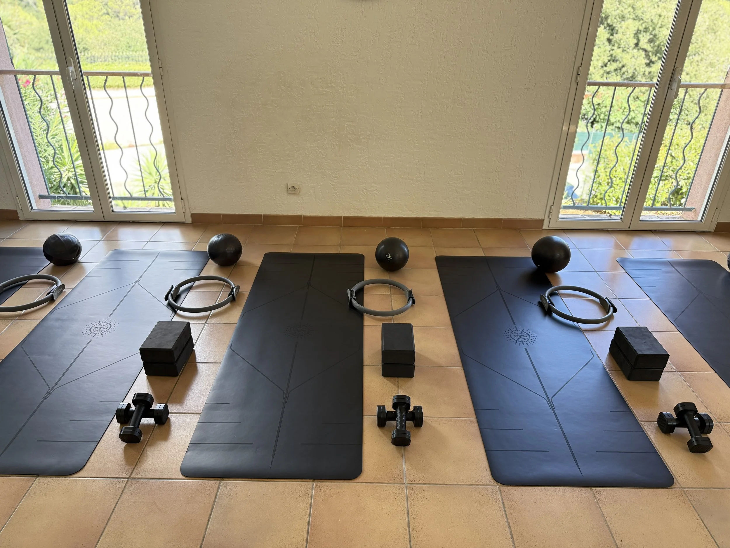 Three black yoga mats with workout equipment on them, including balls, blocks, rings, dumbbells, and a small black box, placed on a tiled floor near a glass door with a view of a green outdoor area.