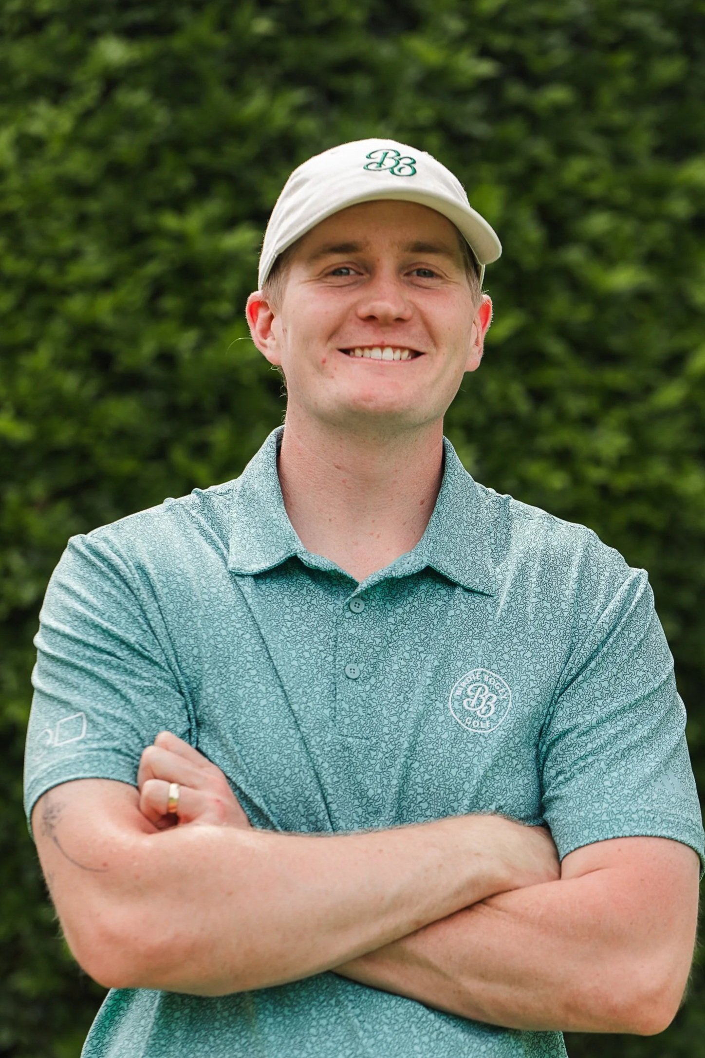 A young man standing outdoors with arms crossed, wearing a white cap and a teal golf shirt, smiling at the camera, with green foliage in the background.