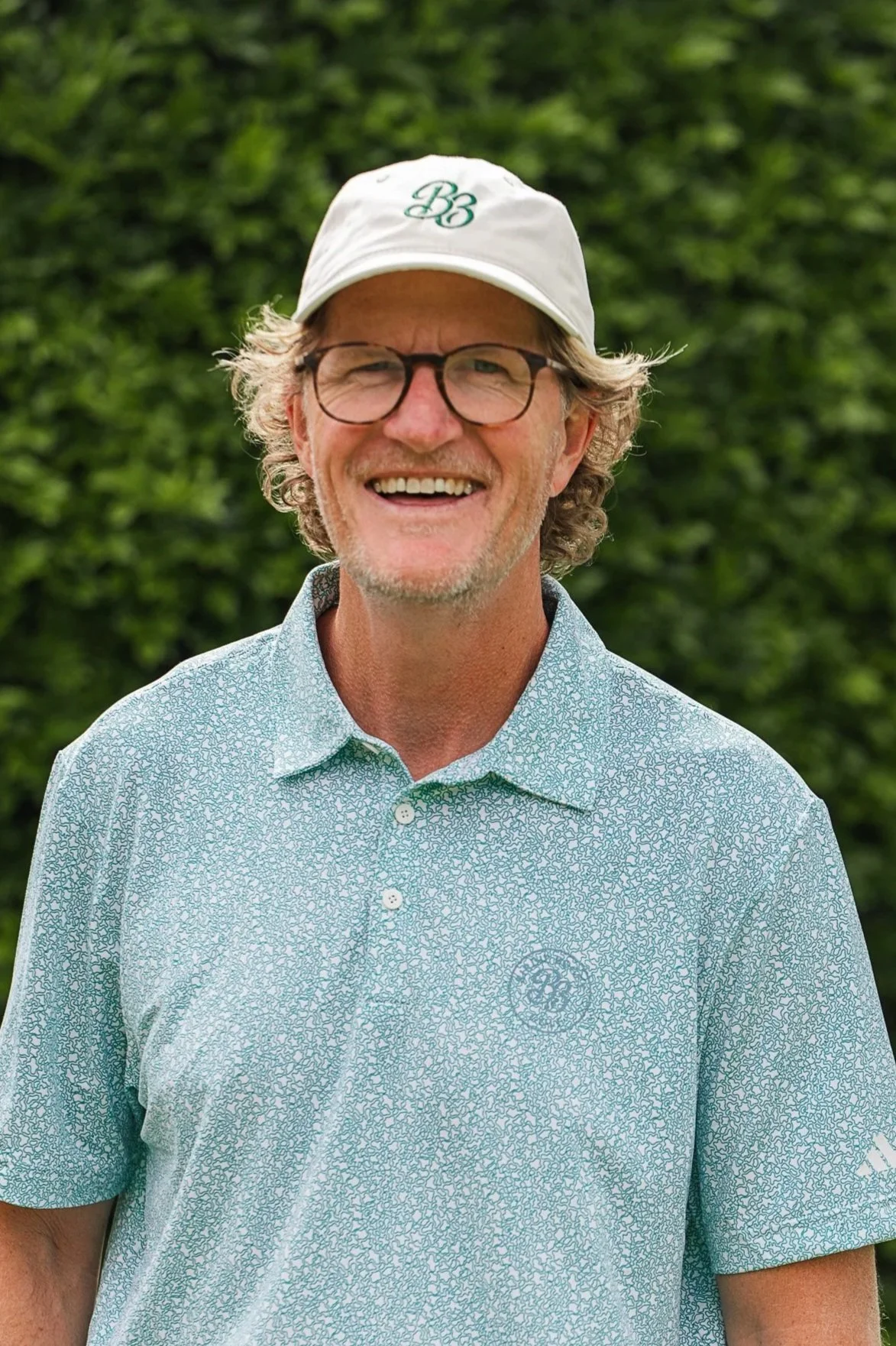 A smiling middle-aged man with curly gray hair wearing glasses, a white cap with a logo, and a patterned golf shirt, standing outdoors with green foliage in the background.