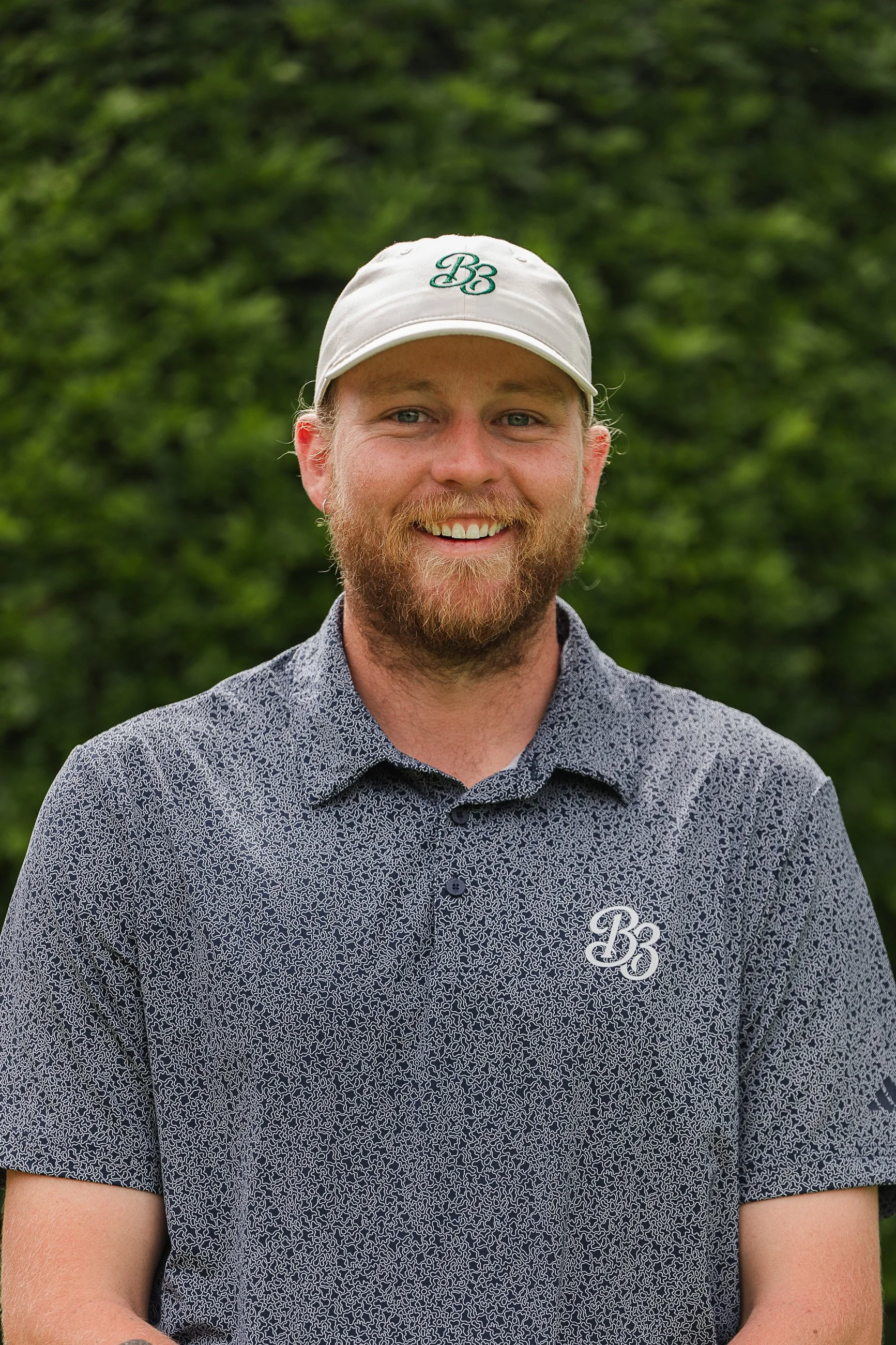 A smiling man with a beard and light hair wearing a white baseball cap with a green embroidered 'B3' and a patterned short-sleeve shirt with a similar logo, standing outdoors with a green, blurred background of trees.