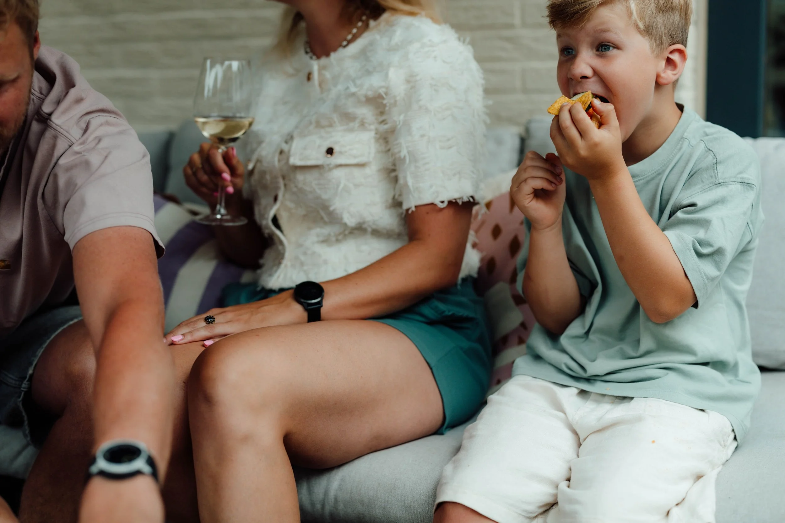 Een bonuskind eet een snack terwijl hij op een bank zit, met zijn moeder en zijn vader naast hem, de vrouw houdt een glas witte wijn vast.