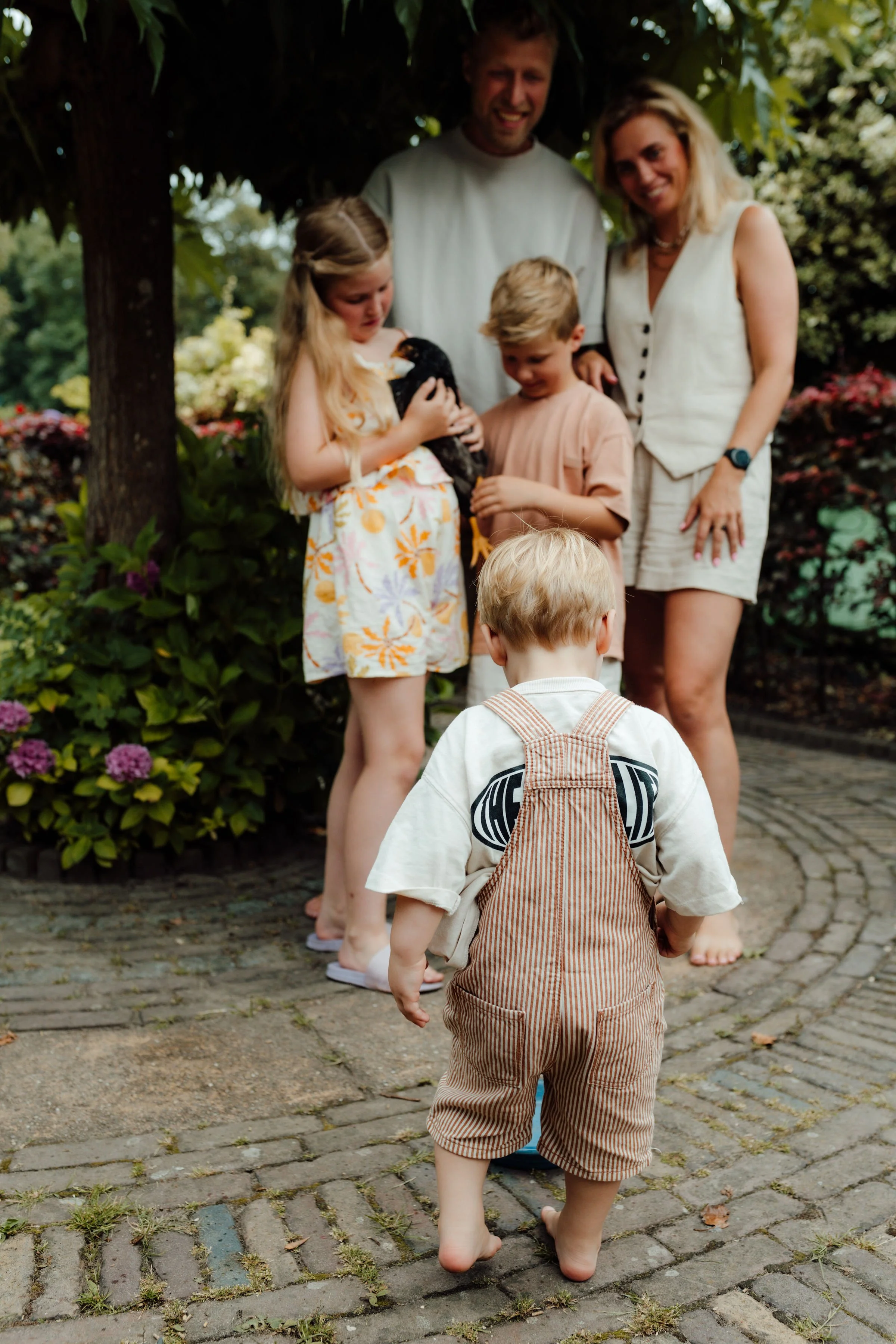 Een samengestelde familie, bestaande uit twee ouders en drie kinderen, staan buiten onder een boom. Eén kind houdt een kleine puppy vast, terwijl de andere kinderen en volwassenen naar de puppy kijken en ervan genieten.